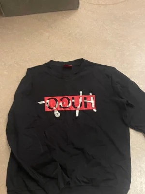 Svart sweatshirt från Hugo Boss - Säljer en svart sweatshirt från Hugo Boss med stor röd och silverfärgad logga framtill. Tröjan har rund halsringning och långa ärmar. Perfekt för dig som vill ha en stilren men ändå statement tröja. Passar till M och S