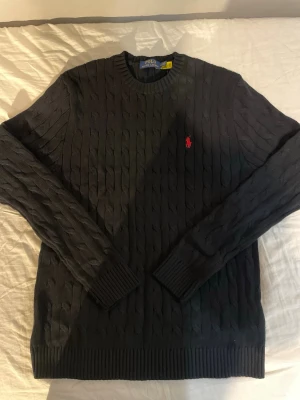 Svart kabelstickad tröja Polo Ralph Lauren - Klassisk svart kabelstickad tröja från Polo Ralph Lauren med rund halsringning och röd broderad logga på bröstet. Tröjan har långa ärmar och ribbade muddar vid ärmslut och nederkant. Perfekt för dig som gillar stilren och tidlös design.