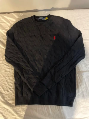 Svart kabelstickad tröja Polo Ralph Lauren - Kabelstickad svart tröja från Polo Ralph Lauren med klassisk röd logga broderad på bröstet. Tröjan har rund halsringning, ribbade muddar och är långärmad. Perfekt för dig som gillar stilrena och tidlösa plagg.