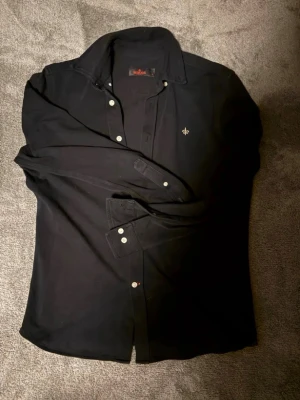 Morris skjorta - Klassisk svart skjorta från Morris med lång ärm och vita knappar. Skjortan har en liten broderad logga på bröstet och är tillverkad i mjuk bomull. Perfekt till jeans eller chinos för en clean look.