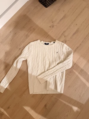 Vit kabelstickad tröja från Gant - Kabelstickad vit tröja från Gant med rund hals och diskret logga på bröstet. Klassisk och stilren design med långa ärmar och ribbade muddar. Perfekt för dig som gillar preppy och tidlös stil.