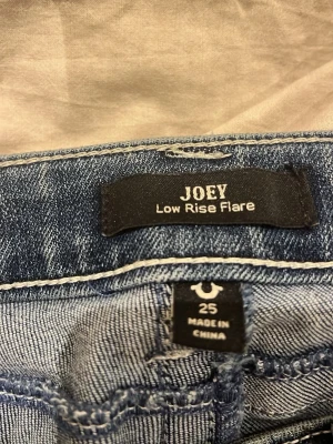 True Religion Joey Low Rise Flare Jeans - Säljer ett par blå True Religion Joey jeans med låg midja och flare-ben. Klassiska kontrastsömmar och stora fickor med lock och dekorativa knappar bak. Tillverkade i jeansmaterial med snygg tvätt och detaljerad sömnad. Har bara använt ett par få gånger.