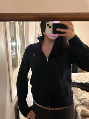 Mörkblå stickad kofta från Ralph Lauren - Säljer en mörkblå stickad kofta från Ralph Lauren med dragkedja framtill och ribbad hög krage. Klassisk RL-logga broderad på bröstet och ribbade muddar vid ärmslut och nederkant. Perfekt till lager på lager under kyliga dagar. Lappen har tyvärr börjat lossna och dragkedjorna är lite missfärgade men mer än det är den i toppen skick