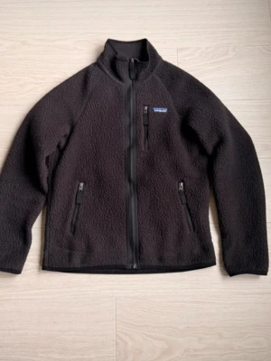 Svart fleecejacka från Patagonia - Mysig svart fleecejacka från Patagonia med hög krage och hel dragkedja framtill. Jackan har två sidofickor med dragkedja och en bröstficka. Perfekt för kyliga dagar och passar perfekt till våren.