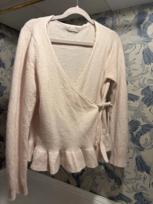 Ljusrosa omlottkofta från Odd Molly med volangkant - Supersöt ljusrosa omlottkofta från Odd Molly med knytband i sidan och volangkant nedtill. Koftan är långärmad och har en mjuk, stickad känsla som passar perfekt till lager på lager. Perfekt för dig som gillar romantiska detaljer och en feminin look.