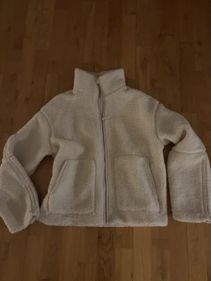 Beige teddyjacka från H&M - Mysig beige teddyjacka från H&M med hög krage och dragkedja framtill. Jackan har två stora fickor och är gjord i fluffigt teddy-material som håller dig varm. Perfekt för kyliga dagar och enkel att matcha med olika outfits. Storlek 164 så passar en xxs/xs