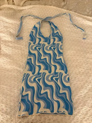 Blåvit kortklänning med vågmönster - Säljer en superfin kortklänning med blått och vitt vågigt mönster. Klänningen har halterneck med knyt i nacken och är ärmlös. Materialet känns som stretchig syntet, perfekt för sommarens alla tillfällen. Snygg och unik design som verkligen sticker ut! Aldrig använd 🥰