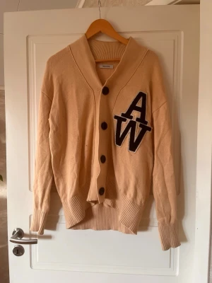 Beige kofta från Jack and Jones  - Säljer en beige kofta från Jack and Jones med stora svarta bokstäver 'AW' på bröstet. Koftan har grova ribbade muddar, stora svarta knappar och en avslappnad passform. Perfekt för lager på lager under höst och vår.