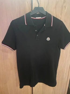 Svart Moncler piké med rödvit kant - Säljer en svart pikétröja från Moncler i storlek S. Pikén har klassisk krage och knäppning framtill samt Moncler-logga på bröstet. Ärmar och krage har snygga rödvita ränder som ger en sportig touch. Tillverkad i bomull för skön känsla. Köpt på NK för 2999kr nu säljs denna piké för 999kr. Nyskick, alltså använd 1 gång på grund av att den är för liten. Perfekt nu inför de varmare tiderna.