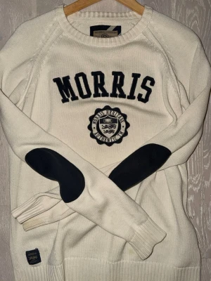 Vit stickad tröja från Morris storlek M - Säljer en vit stickad tröja från Morris med svarta detaljer och broderad logga på bröstet. Tröjan har svarta armbågslappar och ribbade muddar vid ärmslut och nederkant. Perfekt för dig som gillar klassisk college-stil.