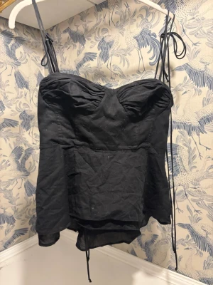 Svart korsettopp med knytband - Säljer en svart korsettopp med tunna axelband som knyts på axlarna. Toppen har en lätt utsvängd nederdel och rynkade detaljer vid bysten. Supersöt topp från Zara som går att justera i banden 