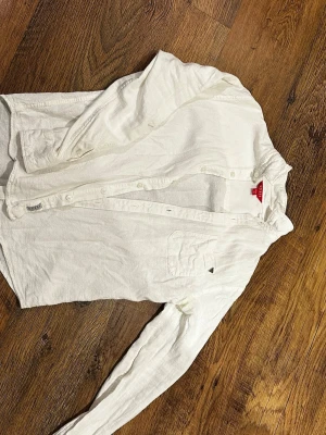 Vit skjorta från Guess XS - Säljer en stilren vit skjorta från Guess i storlek XS. Skjortan har klassisk krage, bröstficka med metalltriangel och knappar framtill. Tillverkad i mjuk bomull som känns skön mot huden. Perfekt till jeans för en clean look.