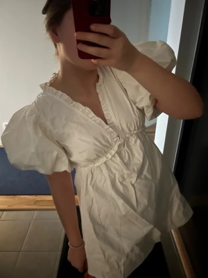 Vit kort studentklänning med puffärmar - Supersöt vit kort klänning med djup v-ringning och volangkant. Klänningen har puffiga korta ärmar och markerad midja. Perfekt för sommarens alla tillfällen och ger en romantisk vibe. Materialet känns lätt och luftigt.