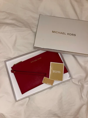 Röd kuvertväska från Michael Kors - Säljer en snygg röd kuvertväska från Michael Kors med guldfärgade detaljer och dragkedja. Aldrig använd, i perfekt skick. 