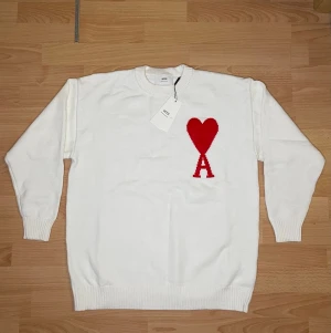 Vit stickad tröja från Ami Paris - Säljer en vit stickad tröja från Ami Paris med rött hjärta och bokstaven A broderat på bröstet. Tröjan har rund hals, ribbade muddar och är långärmad. Perfekt för dig som gillar stilrena plagg med en twist.