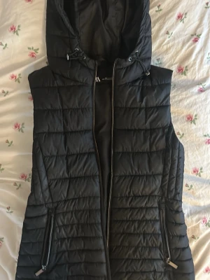 Svart dunväst med huva från Zara - Säljer en svart dunväst från Zara, fickor med dragkedja och quiltad design. Perfekt att slänga över en hoodie för extra värme.