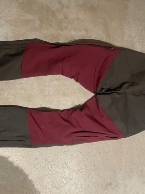 Pinewood vinröda och svarta friluftsbyxor - Säljer ett par Pinewood friluftsbyxor i vinrött och svart. Byxorna har flera dragkedjeförsedda fickor, förstärkta partier och slitstarkt material. Perfekta för uteliv och äventyr. Extra knappar medföljer. Snygg och funktionell design!