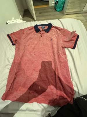 Röd piké från Gant med marinblå krage - Säljer en röd pikétröja från Gant i storlek XL. Skjortan har marinblå krage med röd kant, knappar framtill och diskret Gant-logga broderad på bröstet. Materialet är mjukt och andas, perfekt för sommardagar.