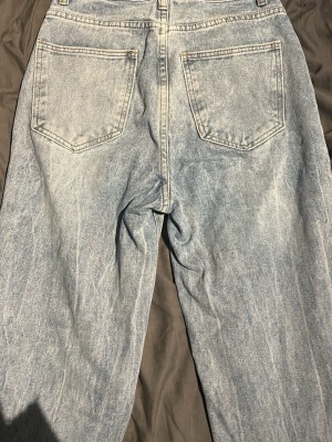Ljusa ripped jeans med raka ben - Säljer ett par ljusblå jeans med raka ben och stora slitna hål vid knäna samt mindre slitningar upptill. Jeansen har klassiska bakfickor och är gjorda i jeansmaterial. Perfekt för dig som gillar en edgy och avslappnad stil.