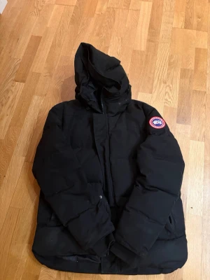 Svart Canada Goose pufferjacka  - Säljer en svart pufferjacka från Canada Goose i storlek M. Jackan har huva, dragkedja och den klassiska loggan på ärmen. Perfekt för kalla dagar och riktigt varm tack vare dunfyllning. Enkel, stilren design som funkar till allt. 