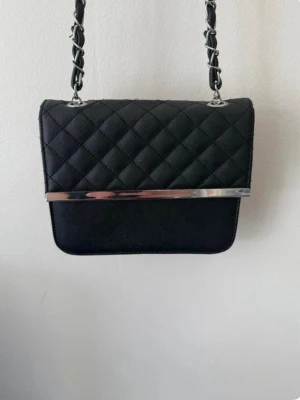 Svart quiltad axelväska med kedja - Snygg svart axelväska med quiltat mönster och silverfärgad kedja i axelbandet. Väskan har en stilren metallist framtill och klassisk rektangulär form. Perfekt för dig som vill ha en trendig och tidlös accessoar.