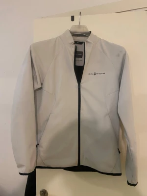 Vit vindjacka från Sail Racing - Säljer en stilren vit vindjacka från Sail Racing i storlek Small. Jackan har svart dragkedja, diskret logga på bröstet och ryggen samt två sidofickor. Materialet är slätt och känns sportigt, perfekt för dig som gillar en clean look.