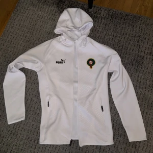 Marocko kofta  - Säljer en vit träningsjacka från Puma med huva och dragkedja. Jackan har ett tryckt emblem på bröstet och två fickor med dragkedja. Perfekt för sportiga aktiviteter dragkedjan är sönder.