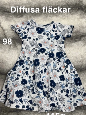 Vit blommig klänning  - Supersöt vit klänning med blått och rosa blommönster. Klänningen har korta ärmar och en mjuk, snurrig kjol. Perfekt för varma dagar och lek utomhus. 🌸 Diffusa fläckar. Storlek 98