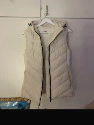 Kräm vit dunväst från Zara med huva - Säljer en kräm vit/ vit dunväst från Zara i storlek M. Västen har huva, svarta detaljer vid dragkedjan. Aldrig använt den för den va inte riktigt min stil. Är i storlek M men jag har S och den passar mig också.