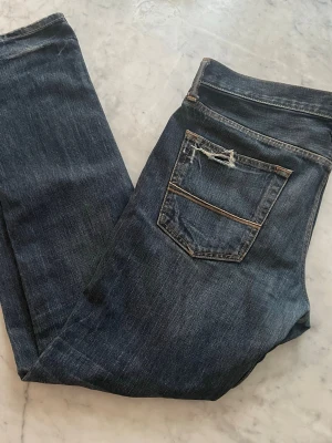 Abercrombie & Fitch blå jeans, straight - Säljer ett par blå jeans från Abercrombie & Fitch med slitna detaljer och trasiga partier på både fram- och baksida. Jeansen har klassisk femficksdesign, raka ben och orange kontrastsömmar. Materialet är jeans i bomull och passformen är straight/slim.