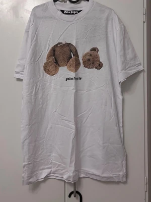  vit t-shirt med björntryck - Säljer en clean vit t-shirt  i storlek XXL passar XL oxå. Framsidan har ett stort tryck med en ikonisk nalle i brunt och svart, där huvudet är separerat från kroppen. Klassisk rund hals och korta ärmar. Materialet är mjuk bomull och loggan syns under trycket.