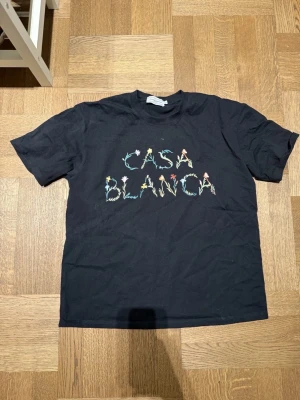 Svart Casablanca t-shirt med blommigt tryck - Snygg svart t-shirt från Casablanca med färgglatt blommigt tryck som stavar 'CASABLANCA' på bröstet. Klassisk passform och rund halsringning. Perfekt för dig som vill ha en unik och trendig tee i garderoben.