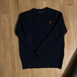 Mörkblå stickad tröja Polo Ralph Lauren - Säljer en mörkblå stickad tröja från Polo Ralph Lauren med klassiskt kabelstickat mönster och röd broderad logga på bröstet. Tröjan har rund hals och långa ärmar med ribbade muddar. Perfekt för dig som gillar stilrena och tidlösa plagg.