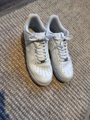 Nike Air Force 1 vita sneakers - Klassiska Nike Air Force 1 sneakers i helvitt skinn med perforerad tå, rund tå och snörning. Tidlös modell med platt sula och diskret Nike-logga på sidan. Perfekta för dig som vill ha en clean och stilren look. Storlek 44
