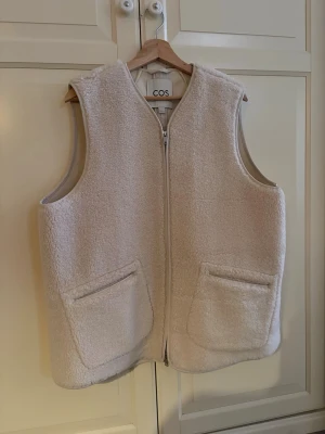 Pileväst - Säljer en krämvit väst i pile från COS i L med dragkedja framtill och två stora fickor. Västen har en rak och loose passform, perfekt att slänga över en hoodie eller tröja. Materialet är mjukt och fluffigt, vilket ger en mysig vibe😊endast använd två gånger så så gott som ny!