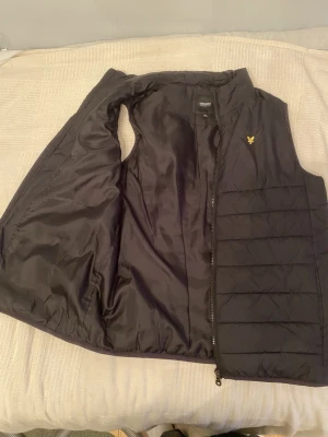Svart dunväst från Lyle & Scott herr  - Säljer en svart dunväst från Lyle & Scott Junior med gul logga på bröstet. Perfekt till våren. Västen har fet design, hög krage och dragkedja framtill. Storlek 15/16 år. Jag på bilden är 186 cm och den är lite kort för mig. Finns även en rymlig innerficka på höger sida dragkedjan framifrån.