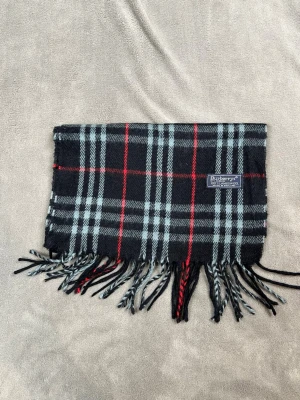 Rutig ullhalsduk från Burberrys - Snygg halsduk från Burberrys i 100% lammull med klassiskt rutigt mönster i svart, grått och rött. Halsduken har tvinnade fransar i ändarna och en vävd logotypetikett. Perfekt accessoar för att lyfta din vinterstil. P9