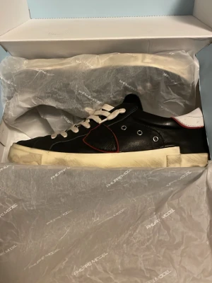 Svarta Philippe Model sneakers i skinn - Säljer ett par svarta Philippe Model sneakers stl 41 i skinn med vita snören och vit sula. Skorna har en vit häl med röd detalj och metalliska öljetter på sidan. Skorna är använda en sommar ungefär och är i bra skick, ända tecken på användning egentligen är på hälen vilket kan ses på bild pågrund av häldrag när jag gått med dem. Skriv gärna om ni har några frågor eller vill ha fler bilder!😊Skorna kommer i originalbilden. Pris kan diskuteras vid snabb och enkel affär.