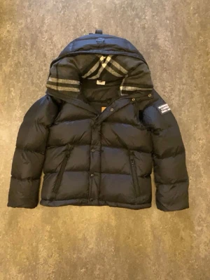 Svart pufferjacka från Burberry - Svart pufferjacka från Burberry med avtagbara ärmar så du kan bära den som väst. Jackan har dragkedja och knappar framtill, två fickor med dragkedja och en huva med klassiskt rutigt Burberry-mönster på insidan. Perfekt för kalla dagar och riktigt snygg stil.