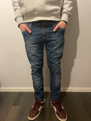 Blå jeans från Nudie Jeansco W29 L32 - Snygga blå jeans från Nudie Jeans i klassisk modell med raka ben och normal passform. Jeansen har kontrastsömmar, fem fickor och coola detaljer på bakfickorna. Perfekt för dig som gillar stilrena och tidlösa jeans med lite edge.