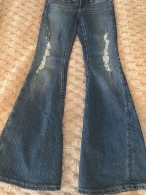 Blå bootcut jeans Gina Tricot XS 14+ - Säljer ett par blå bootcut jeans från Gina Tricot i storlek XS. Jeansen har slitna detaljer framtill, klassisk femficksmodell och breda ben. Materialet är jeans med snygg tvätt och kontrastsömmar. Perfekta för dig som gillar retrovibbar och statementbyxor.