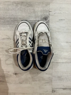 Vintage Adidas Sneakers, Vit & Blå - Y2K Estetik - Snygga Adidas sneakers i vitt och mörkblått med en rejäl, retroinspirerad design. Original priset var 1000kr. Pris kan självklart diskuteras   Skorna har den klassiska designen med tre ränder och en tjock sula som ger bra dämpning och komfort. Stilen påminner om populära modeller från sent 90-tal och tidigt 2000-tal, som till exempel Response CL-serien eller Eclyptix 2000. Stl: 37/38  Skorna är begagnade men i bra skick, perfekta för att komplettera en streetwear eller vintage-inspirerad look. 
