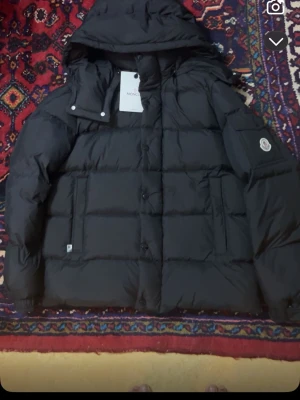 Svart Moncler dunjacka med huva - Säljer en svart Moncler dunjacka med huva och stora fickor. Jackan har quiltad design, dragkedja och knappar framtill samt Moncler-logga på ärmen. Perfekt för kalla dagar och riktigt snygg till streetwear-stilen.