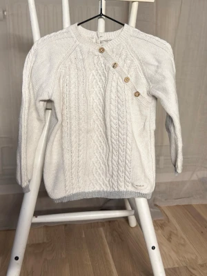 Vit stickad kofta från Newbie - Supersöt vit stickad kofta från Newbie med flätmönster framtill och träknappar i sned knäppning. Långärmad modell med ribbade muddar och grå detaljer på armbågar och nederkant. Perfekt för kyliga dagar!