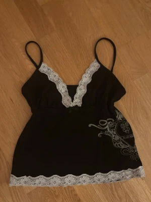 Svart spetslinnetopp från SHEIN  - Säljer en svart linnetopp från SHEIN med tunna axelband och dekorativ vit spets längs ringningen och nederkanten. Toppen har en liten rosett framtill och ett tryckt mönster i grått på sidan. Perfekt för dig som gillar detaljer.