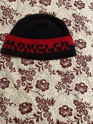 Svart och röd Moncler mössa - Original snygg svart mössa från Moncler med bred röd kant och stor logotyp framtill. Mössan har en klassisk form och är tillverkad i mjukt material som sitter skönt på huvudet. Perfekt för dig som gillar streetwear och vill ha en cool accessoar med tydlig branding.