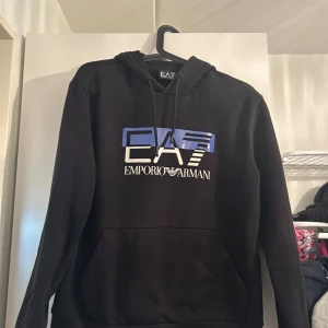 Svart EA7 Emporio Armani hoodie - Svart hoodie från EA7 Emporio Armani med stor logga i vitt och blått framtill. Tröjan har huva med snörning och en klassisk känguruficka. Perfekt för dig som gillar stilrena streetwear-plagg med premiumkänsla.