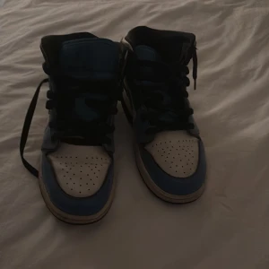 Nike Air Jordan 1 blå och vita sneakers - Säljer ett par Nike Air Jordan 1 sneakers i blått och vitt med svarta detaljer. Skorna har klassisk snörning, svart sula och perforerad tåbox. Ikonisk design med högt skaft och tydlig Jumpman-logga på plösen. Perfekta för dig som gillar streetwear och vill sticka ut. INGEN BOX BARA SKORNA 