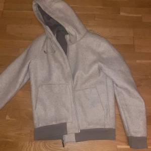 Ljusgrå hoodiejacka från Dan John - Snygg baige hoodiejacka från Dan John med dragkedja, stora fickor framtill och ribbade muddar i mörkare grått. Jackan har huva med snörning och är tillverkad i ett mjukt material som passar perfekt för kyligare dagar. Tröjan är gjord och köpt I Italien/Rom i en lyxig affär. Denna jacka är perfekt till lite kallare dagar nu på hösten.