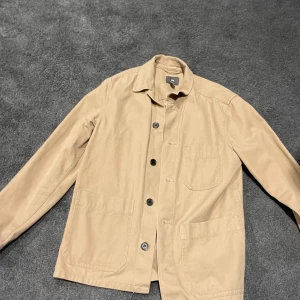 Beige overshirt från H&M - Stilren beige overshirt från H&M med klassisk krage och tre stora fickor framtill. Jackan har knäppning med svarta knappar och är tillverkad i ett mjukt bomullsmaterial. Perfekt för lager-på-lager och enkel att matcha med olika outfits.